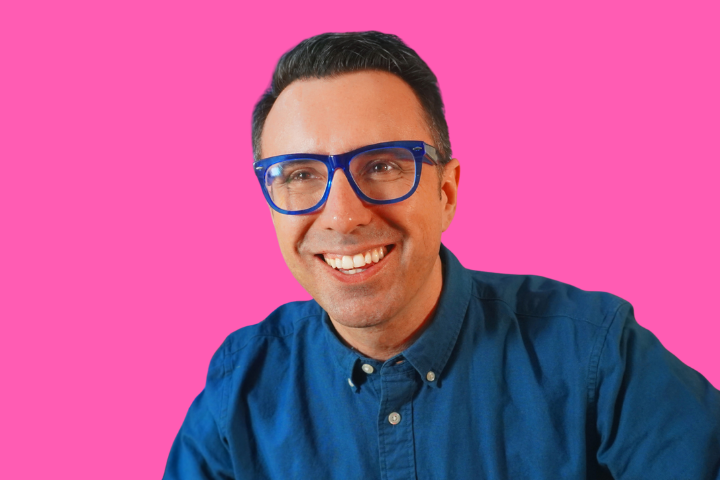 A smiling man wearing bright blue glasses and a blue button-up shirt stands in front of a solid pink background.
