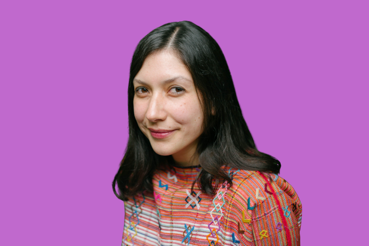 A woman with long dark hair, wearing a colorful patterned top, smiles slightly while posing in front of a solid purple background.