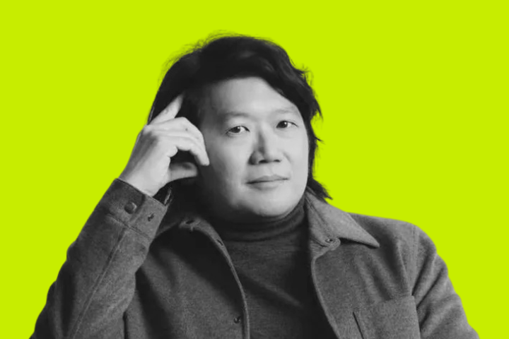 A person with medium-length dark hair, wearing a turtleneck and coat, sits with one finger resting on their temple against a bright yellow background.