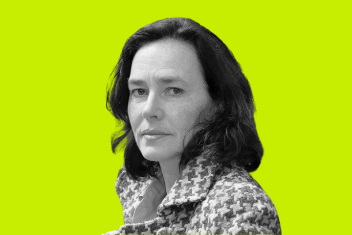 A woman with dark wavy hair, wearing a houndstooth-patterned coat, looks at the camera. The background is a solid bright yellow-green color. The image is in black and white except for the background.