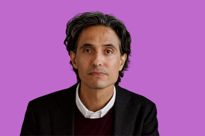 A man with medium-length dark hair and gray streaks, wearing a black blazer over a dark sweater and white shirt, sits against a plain purple background, looking directly at the camera with a neutral expression.