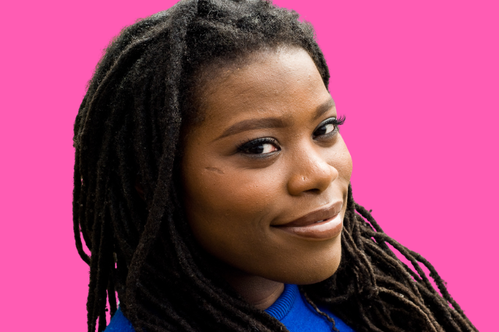 A woman with long dreadlocks smiles slightly while looking at the camera. She is wearing a blue top and is pictured against a solid pink background.