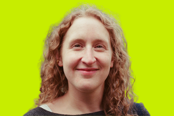 A person with curly, light brown hair smiles at the camera. They are wearing a dark sweater over a light shirt. The background is a solid bright yellow.