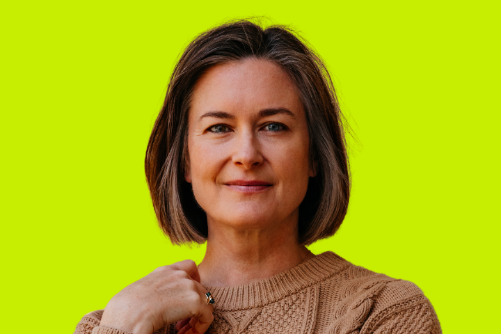 A woman with short brown hair, wearing a tan knit sweater, stands in front of a bright yellow-green background, smiling softly and looking confidently at the camera.