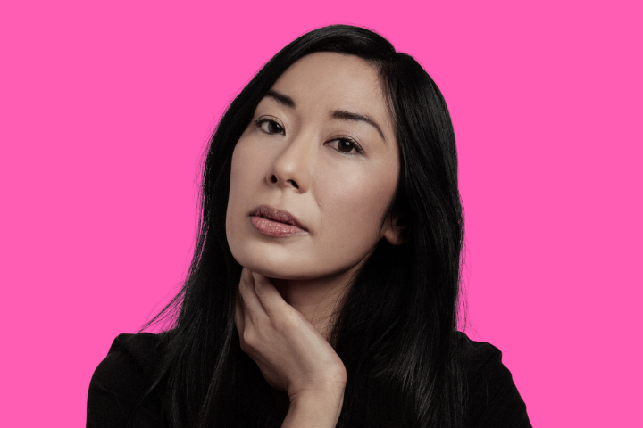A person with long, straight black hair and a neutral expression rests their hand on their neck, posed against a bright pink background.