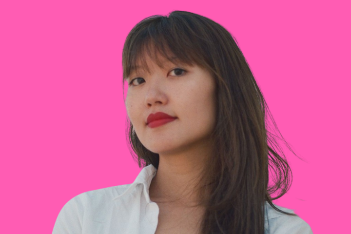 A person with long dark hair and red lipstick wearing a white shirt, standing against a bright pink background, looking confidently at the camera.