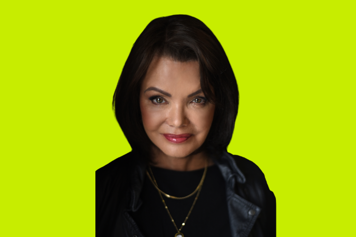 A woman with dark hair and blue eyes, wearing a black jacket and layered gold necklaces, is smiling softly against a bright yellow-green background.