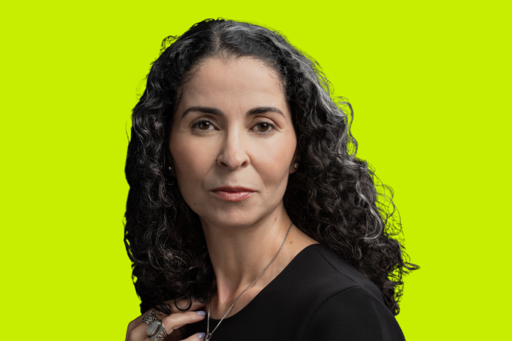 A woman with long, curly dark hair and a serious expression poses against a bright yellow-green background, wearing a black top and silver jewelry.