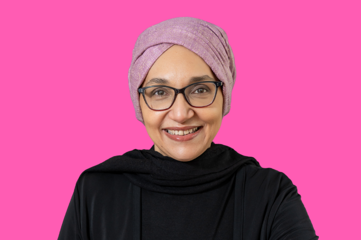 A woman wearing glasses, a light purple headscarf, and a black top smiles at the camera in front of a bright pink background.