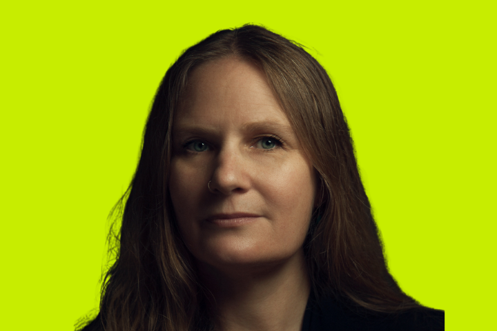 A woman with long brown hair and a neutral expression is shown against a bright yellow-green background. She is facing slightly to the side, wearing a dark top.