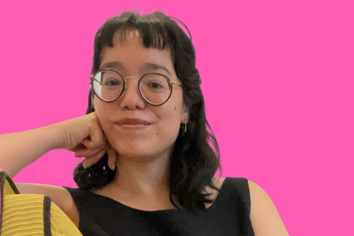 A person with shoulder-length dark hair, glasses, and a black sleeveless top is smiling softly, resting their chin on their hand, sitting in front of a bright pink background.