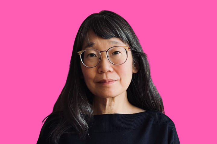 A woman with long dark hair and glasses, wearing a black top, stands in front of a solid pink background.