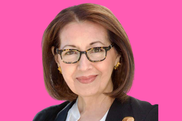 A woman with shoulder-length brown hair, glasses, and gold earrings is smiling. She is wearing a black blazer over a white top, and the background is solid bright pink.