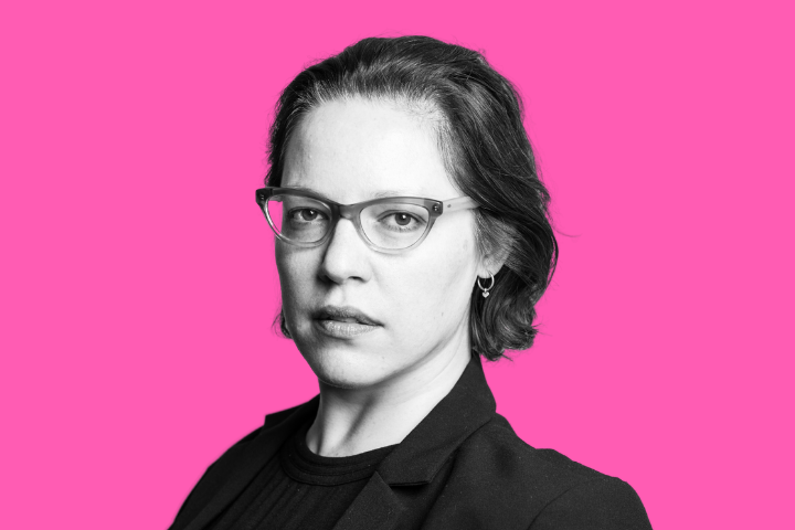 A person with short dark hair, wearing glasses, a dark top, and a blazer looks at the camera against a bright pink background.