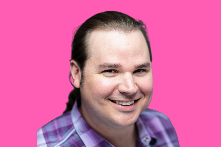 A smiling person with long brown hair tied back, wearing a purple plaid shirt, is pictured against a bright pink background.