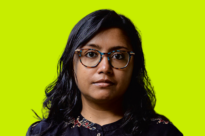 A person with long black hair and glasses, wearing a dark shirt with a subtle pattern, stands in front of a bright yellow-green background, looking directly at the camera with a neutral expression.