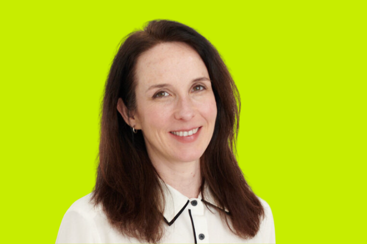 A woman with long dark brown hair, wearing a white collared shirt with black trim, smiles softly in front of a bright yellow-green background.