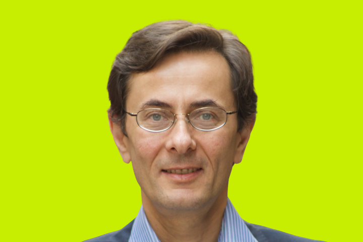 A man with short brown hair, glasses, and a slight smile is wearing a blue collared shirt and dark jacket, posing in front of a bright yellow-green background.