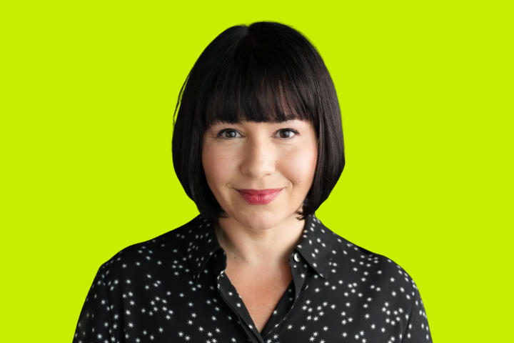 A woman with straight black hair and bangs, wearing a black shirt with white star patterns, smiles in front of a bright lime green background.