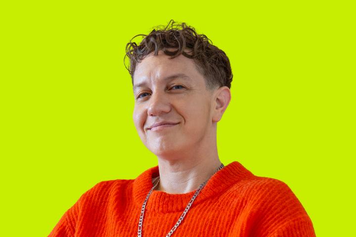 A person with short, curly hair wearing a bright orange sweater and a chain necklace smiles confidently against a vibrant yellow-green background.