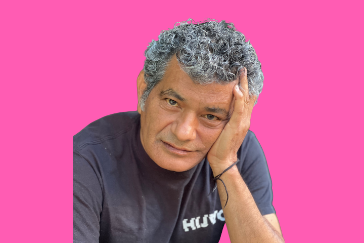 A man with curly gray hair wearing a black T-shirt rests his face on his hand, looking at the camera against a bright pink background.