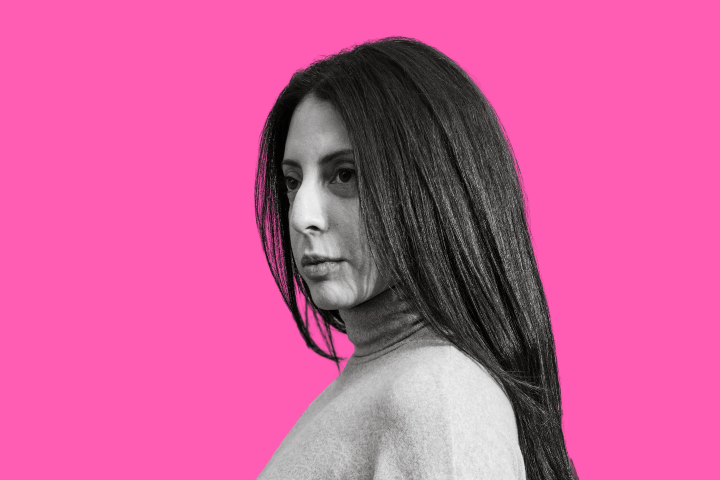 A woman with long, straight dark hair in a turtleneck sweater looks to the side against a bright pink background.