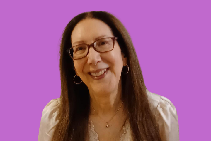 A woman with long brown hair, glasses, and hoop earrings smiles at the camera. She wears a white blouse and a necklace, with a solid purple background behind her.