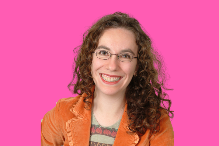 A smiling person with curly brown hair, glasses, and an orange jacket sits in front of a bright pink background.