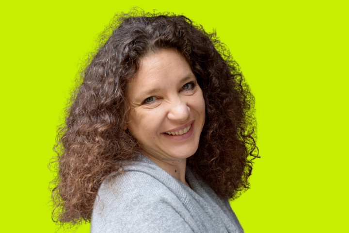A woman with curly brown hair wearing a gray sweater smiles while looking over her shoulder, set against a bright yellow-green background.
