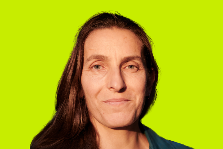 A woman with long brown hair and a faint smile is facing the camera. She is wearing a teal top, and the background is bright yellow-green.