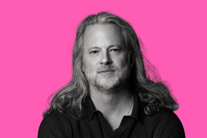A man with long, wavy hair and a beard, wearing a dark collared shirt, sits against a bright pink background, looking directly at the camera with a slight smile.