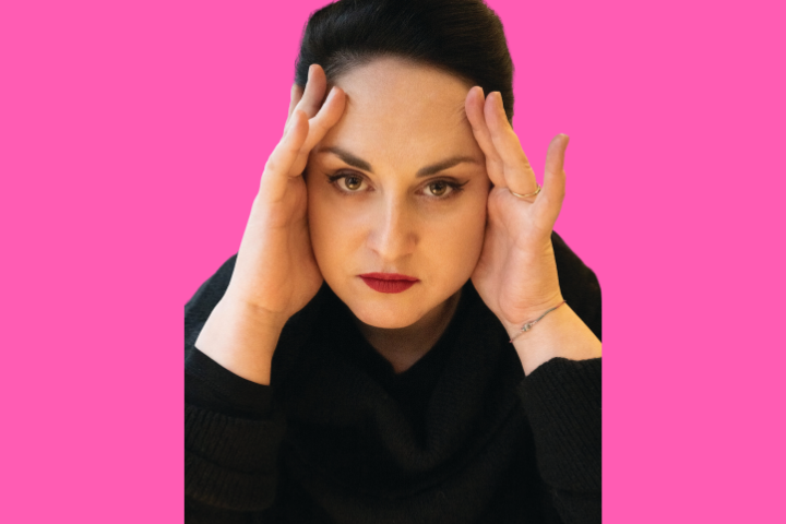 A woman with dark hair pulled back, wearing a black top, rests her hands on her temples and looks forward with a serious expression against a bright pink background.