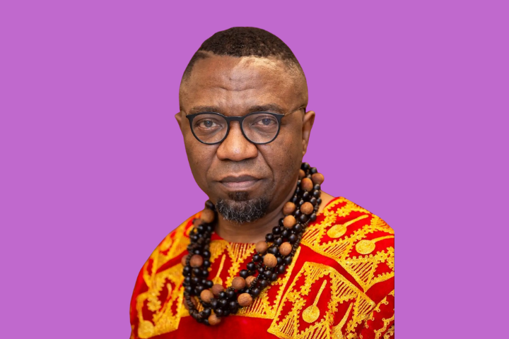 A man wearing glasses, a red and yellow patterned traditional outfit, and layered beaded necklaces poses against a solid purple background.