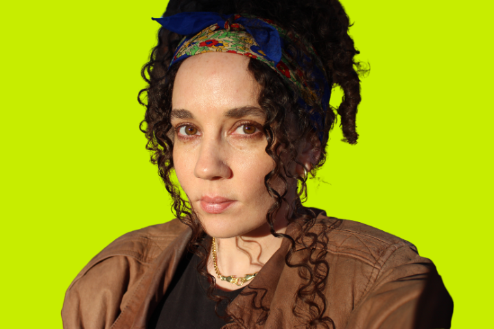 A woman with curly dark hair, wearing a floral headband, brown jacket, and a black shirt, looks seriously at the camera against a bright yellow-green background.