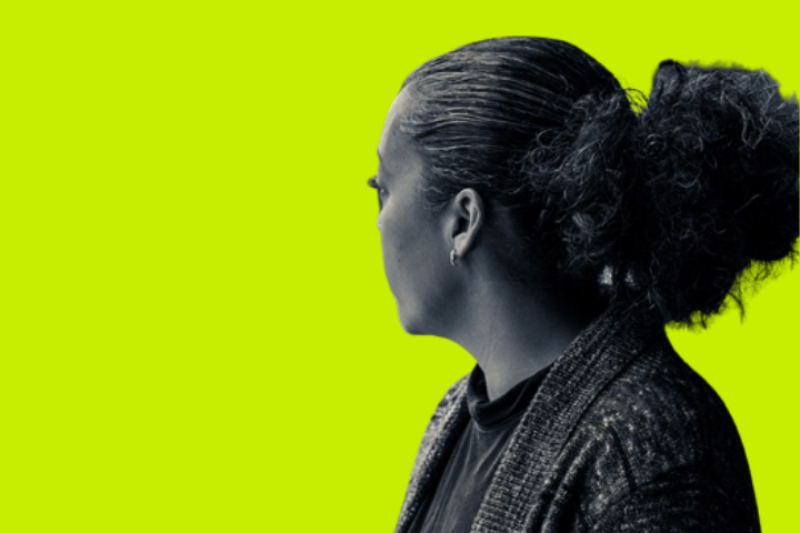 A woman with curly hair in a ponytail, wearing a dark top and cardigan, is facing away from the camera against a bright yellow-green background.