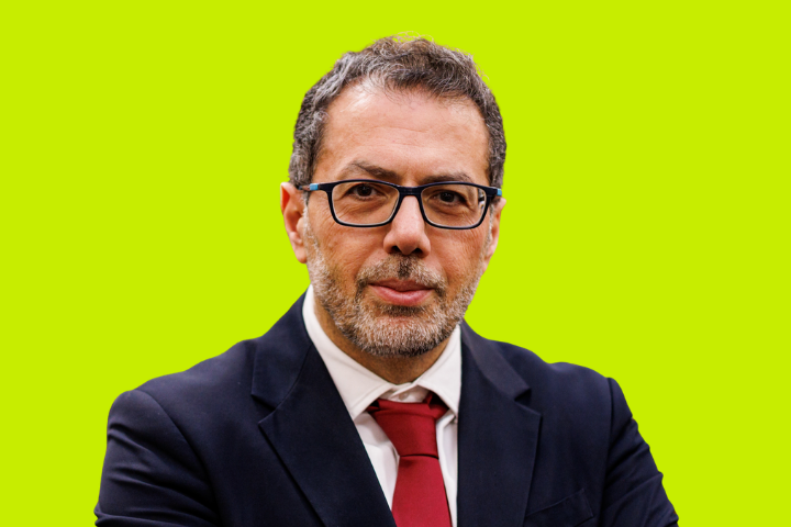 A man with short dark hair, glasses, and a trimmed beard wearing a black suit, white shirt, and red tie, stands against a bright yellow-green background.