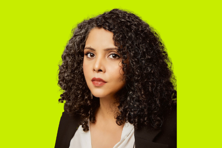 A woman with curly dark hair and a neutral expression, wearing a white top and black blazer, is posed against a bright yellow-green background.
