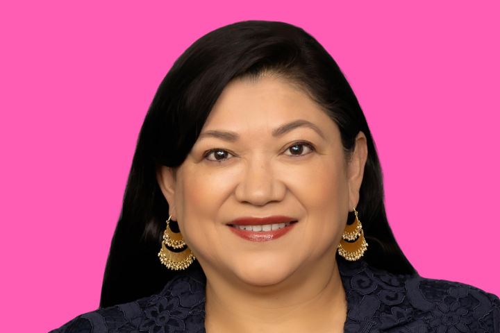 A woman with long dark hair smiles at the camera. She is wearing gold hoop earrings and a dark navy top with a floral pattern, set against a bright pink background.