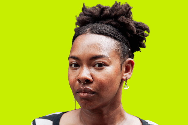 A person with natural curly hair styled up, wearing hoop earrings and a nose ring, looks at the camera with a neutral expression. The background is a bright lime green.