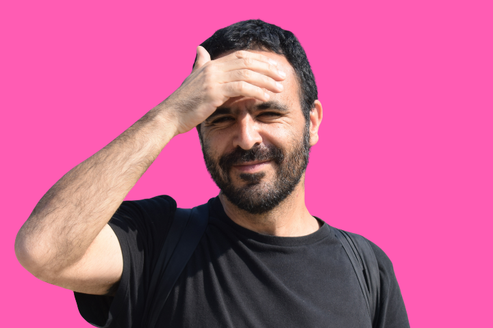A man with a beard and mustache, wearing a black t-shirt and backpack, shades his eyes with his hand while smiling against a solid pink background.