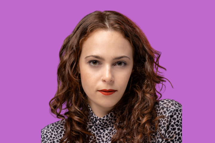 A person with long, curly brown hair and red lipstick wears a leopard-print top, looking directly at the camera against a solid purple background.