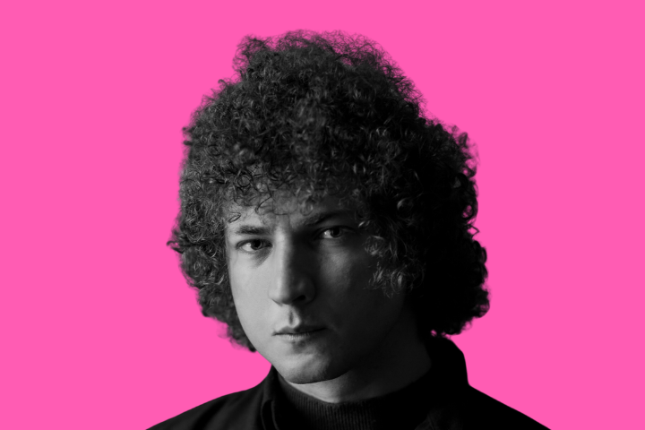 A young man with curly hair and a serious expression is shown in black and white against a bright pink background.