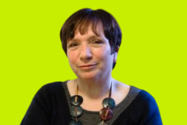 A woman with short brown hair and a neutral expression wears a dark top and a chunky, colorful necklace, posing in front of a bright yellow-green background.