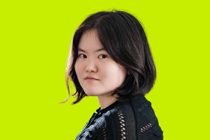 A young person with short, dark hair wearing a black textured sweater looks over their shoulder against a bright yellow-green background.