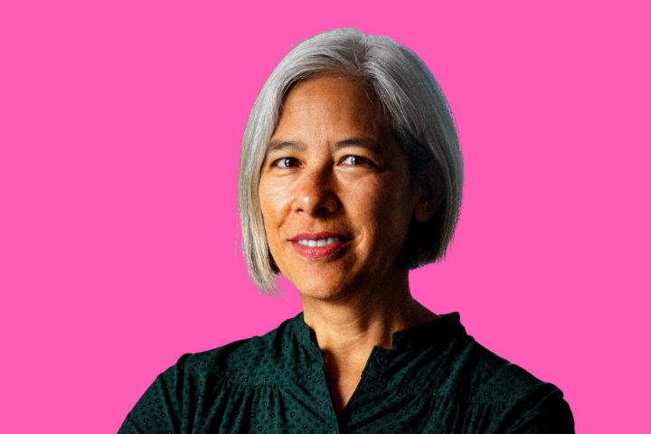 A woman with short gray hair and a dark green blouse smiles confidently in front of a solid bright pink background.
