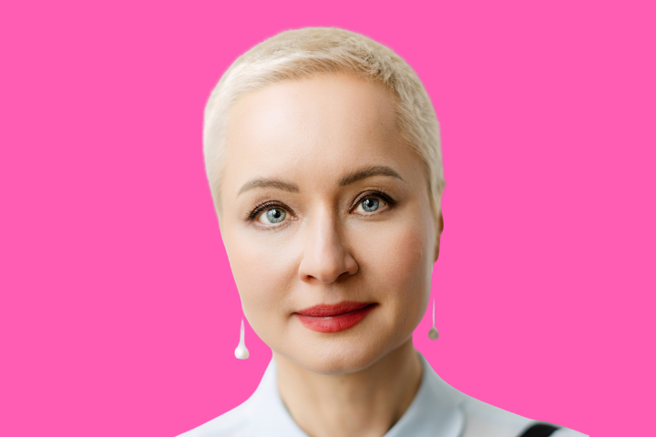 A woman with short platinum blonde hair and light eyes, wearing drop earrings and a light-colored shirt, poses against a bright pink background.