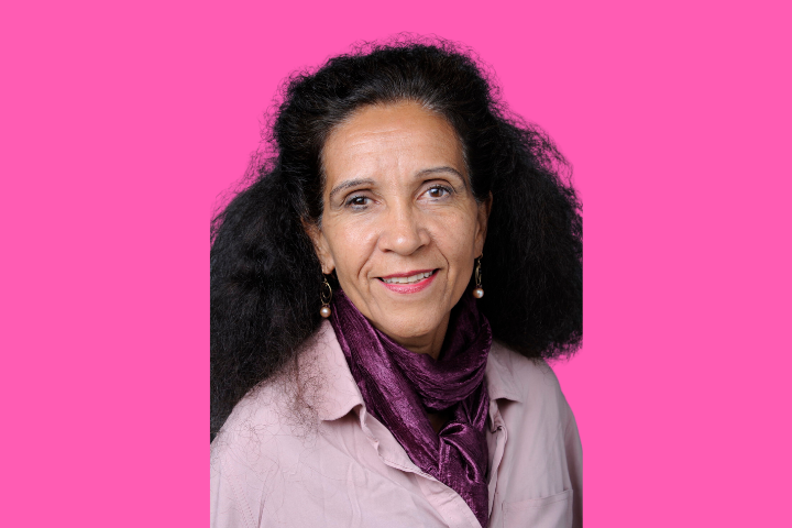 A woman with long, curly dark hair smiles at the camera. She is wearing a pink blouse, a purple scarf, and dangling earrings. The background is bright pink.