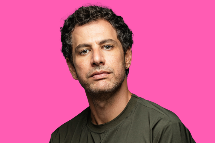 A man with short curly hair, light skin, and slight stubble wears a dark green shirt and small hoop earrings, posing against a bright pink background.