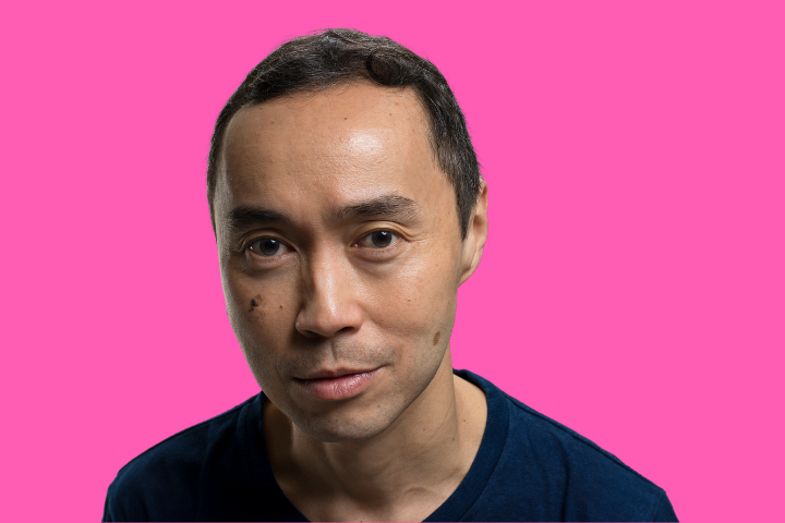 A man with short dark hair and a dark blue shirt looks directly at the camera, standing against a solid bright pink background.