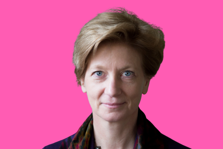 A woman with short light brown hair and blue eyes, wearing a dark jacket and patterned scarf, is looking at the camera against a bright pink background.